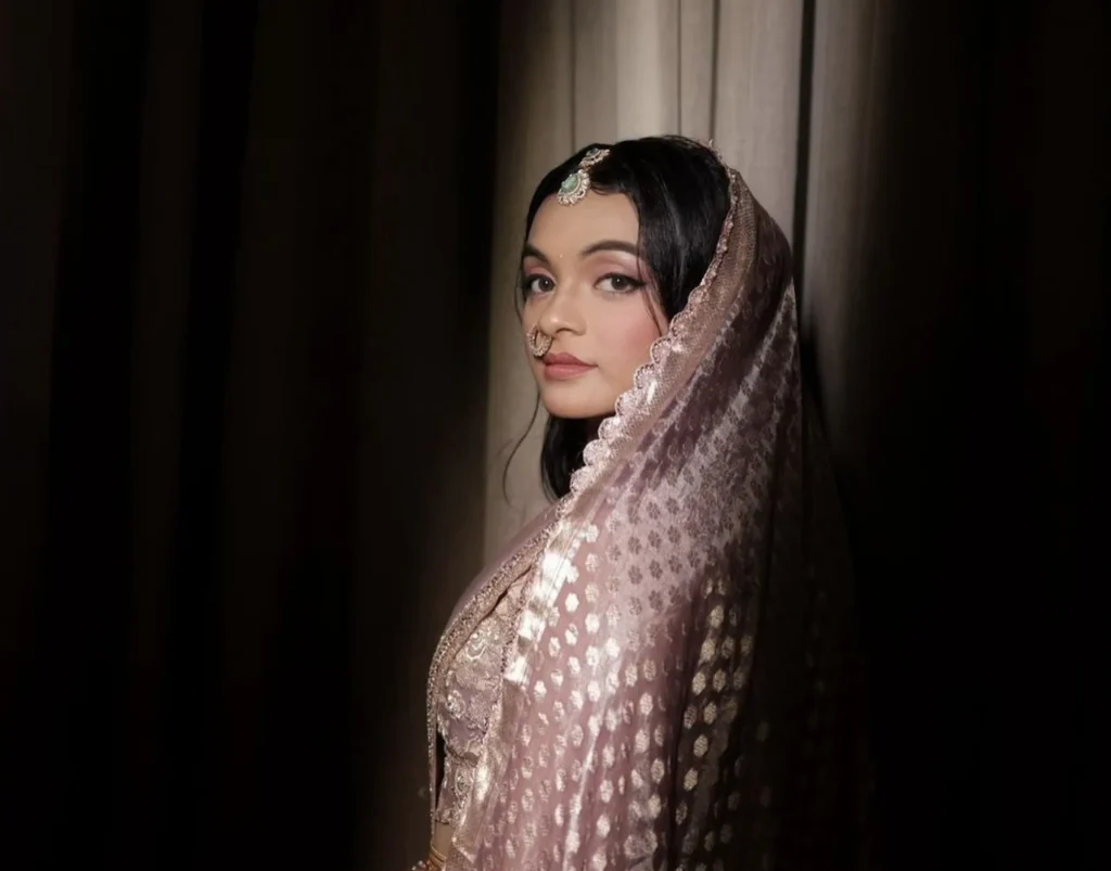 Bride showcasing traditional royal bridal makeup with soft winged eyeliner, a neutral lip, an emerald maang tikka, and a metallic tissue dupatta.